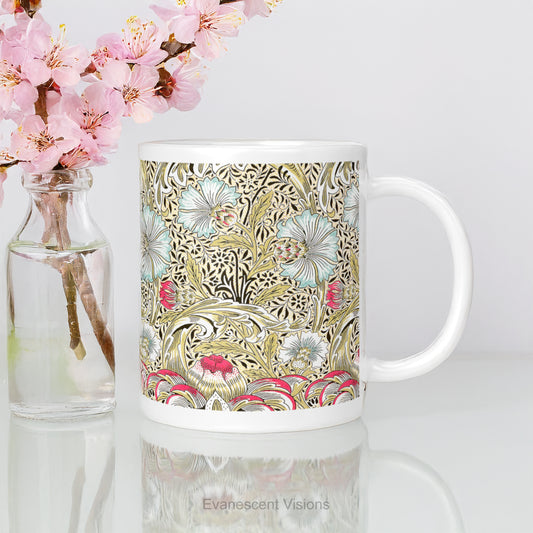 A white ceramic mug featuring the intricate floral design titled 'Corncockle' by William Morris , shown on a white reflective table top with a small vase of pink flowers.