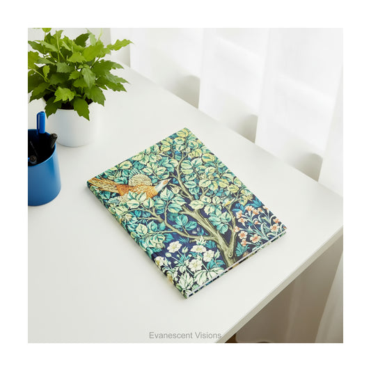 A hardback notebook featuring green Victorian textile design 'Cock Pheasant' by William Morris, lying on a white desk near a window, pen pot and small plant.