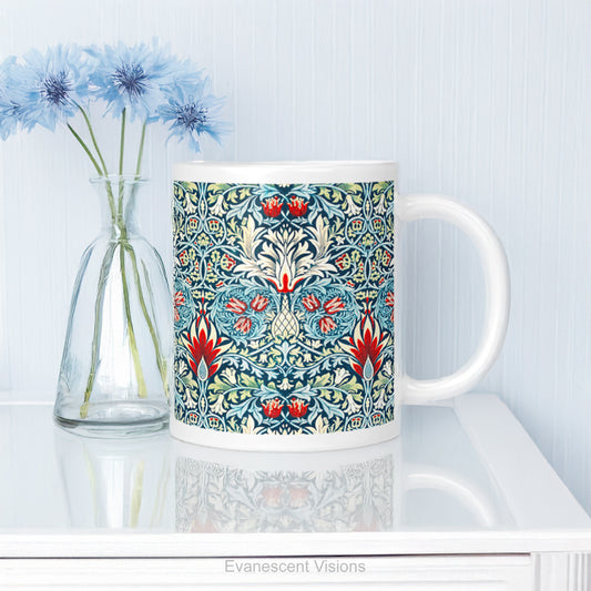 A white ceramic mug featuring a detailed, symmetrical floral pattern in navy blue, sage green, and red called Sankeshead by William Morris, shown on a glass table top.