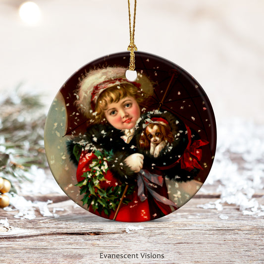 Ceramic Christmas Ornament with design of little girl holding a puppy in a muff in the snow. The ornament rests on a wooden surface with snowflakes and fir tree branch