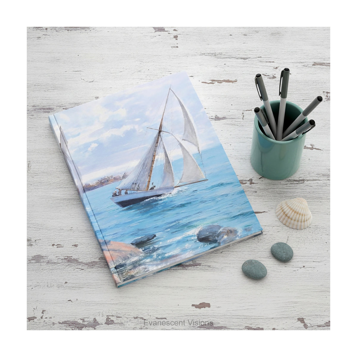 A hardcover notebook  depicting a white sailboat sailing on blue water under a cloudy sky, from painting 'Sailing' by Danielson-Gambogi, shown on a distressed white desk with shells and pebbles. 