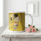 A white ceramic mug featuring a print of  Gustav Klimt's 'The Kiss' painting shown on a white wood table with small pink roses. 