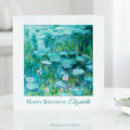 A white greeting card standing on a white surface, featuring Claude Monet's impressionist Water Lilies Painting in blue, green, and teal tones with the text 'Happy Birthday Elizabeth' below the artwork. 