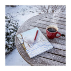 A hardback notebook featuring  a snowy winter landscape, shown on  garden table in the snow.  