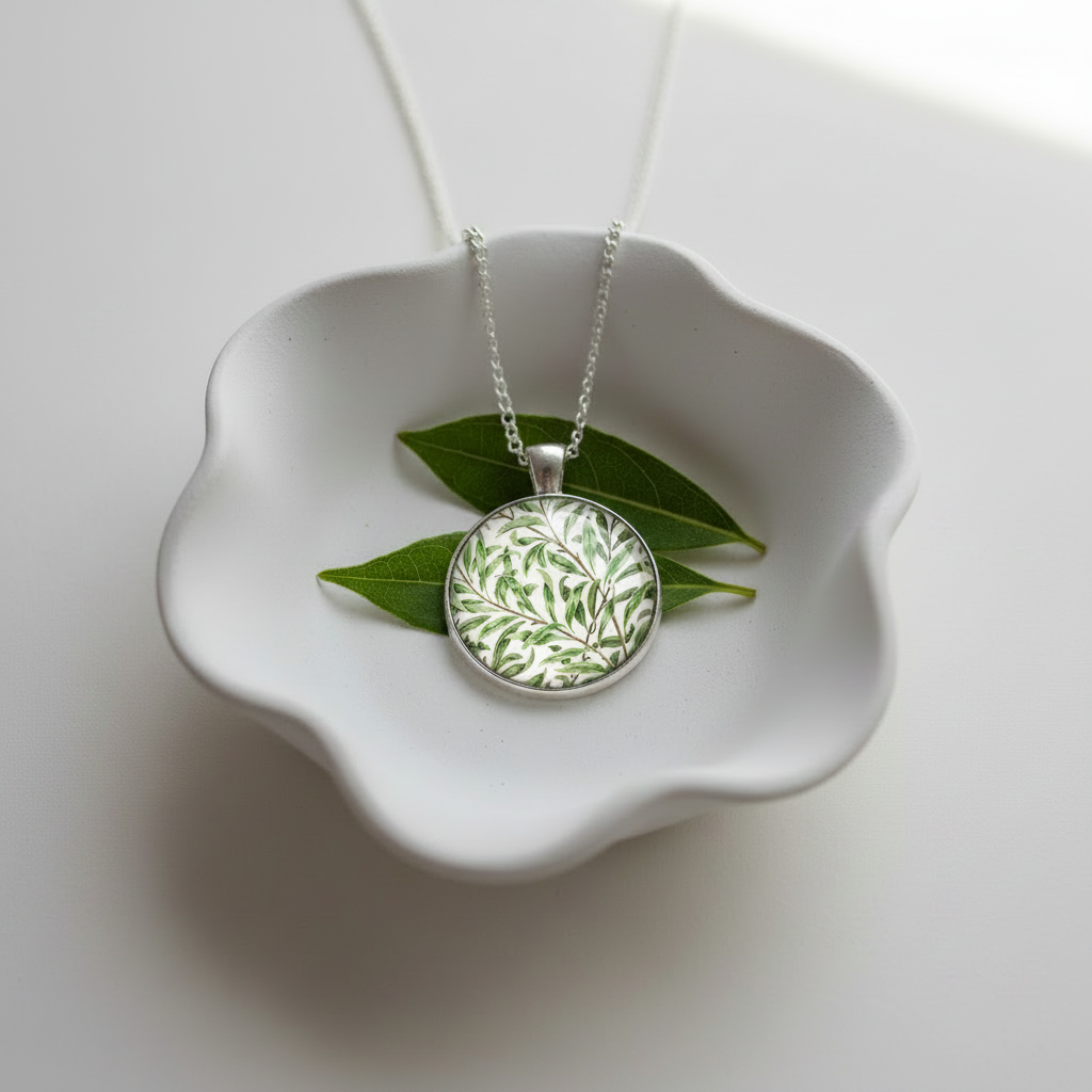 Cabochon pendant necklace with detail from the design  'Willow bough' by William Morris displayed on a white ceramic dish .